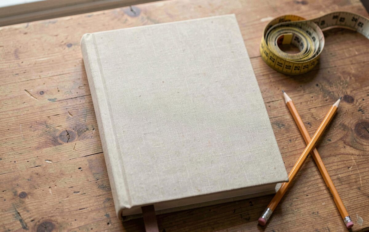 A closed clothbound book on a desk, a tailor's measuring tape coiled beside it